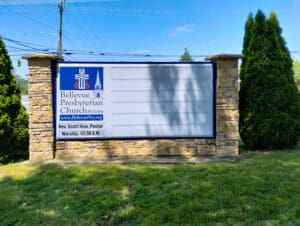 A large white outdoor church sign with blue text, flanked by stone walls and green shrubs, under a clear blue sky.