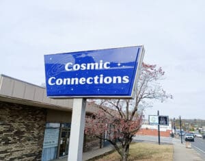 A weathered blue sign reading "cosmic connections" above a commercial building with a partly cloudy sky in the background.