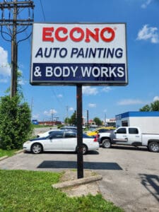 Sign for econo auto painting & body works with parked cars and a clear blue sky in the background.