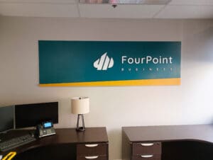 Office wall with "fourpoint business" logo on a teal and orange sign above a desk with a lamp and a telephone.