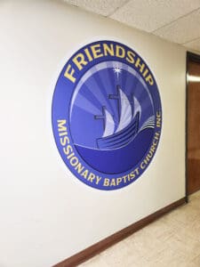 Circular sign of friendship missionary baptist church, featuring a blue background with a white ship symbol, mounted on an interior wall next to a door.