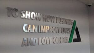 Metallic wall text reads "to show how business can improve lives and love others" with stylized graphics in a corporate setting.