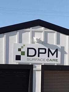 A white cat perched atop a sign reading "dpm surface care" on a building, under clear blue skies.