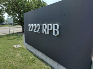 A close-up view of a dark sign with raised silver letters that read "2222 rpb", located near a roadside with greenery in the background.