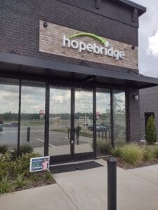 Exterior view of hopebridge center, featuring glass doors, a black facade with "hopebridge" logo, and hiring signs on the door and sidewalk.