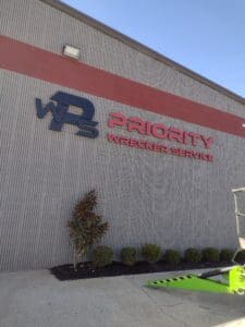 Exterior view of a building with "w3 priority wrecker service" signage, featuring stylized lettering and company logo on a ridged metal facade, under clear blue sky.