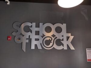 Metallic "school of rock" logo mounted on a gray wall, illuminated by overhead lights, with a red fire alarm box to the left.