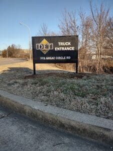 A black sign reading "det, truck entrance, 301 great circle rd" beside a road, with trees in the background and frost on grass.
