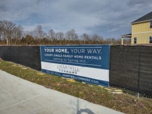 Banner on a construction fence advertising "chartwell commons" luxury single-family rentals, with a background of a clear sky and a house on the right.