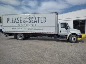 A white delivery truck with "please be seated event rentals" branding on the side, parked at a loading dock under a clear sky.
