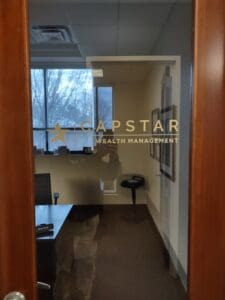 Office door with glass pane displaying "capstar wealth management" logo, leading into a room with a desk and chair, seen from a dimly lit corridor.