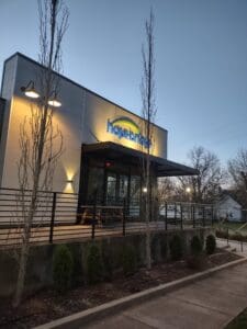 Exterior view of a modern restaurant named "hopdoddy" during dusk, featuring illuminated signage, bare trees, and a pathway leading to the entrance.