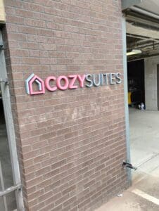 Sign reading "cozysuites" with a stylized house logo, mounted on a brick wall next to an open garage.