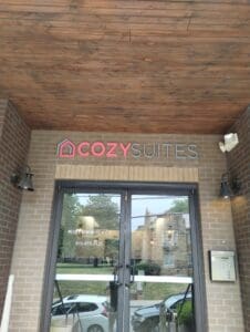 Entrance of "cozy suites" with a logo above glass doors, framed by brick walls and a wooden ceiling, reflecting an outdoor scene.
