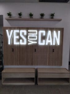 A wall with vertical wooden slats featuring the illuminated letters "yes we can" flanked by benches and topped by a shelf with small plants.