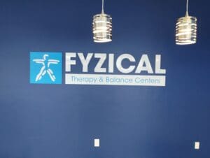 Logo of fyzical therapy & balance centers on a blue wall, flanked by two silver pendant lights.