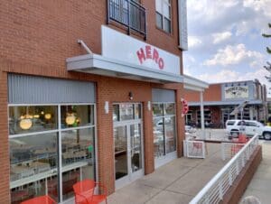 Exterior view of a modern restaurant named "hero" with red and white signage, large windows, and outdoor seating along a city sidewalk.