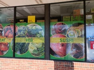 Storefront window display featuring large food images labeled "eat," "veg," and "sushi," with western union and money transfer advertisements.