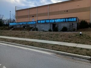 Exterior view of tabernacle baptist church with a banner stating "we want you here sundays 9-9:45am" displayed on the building's side.