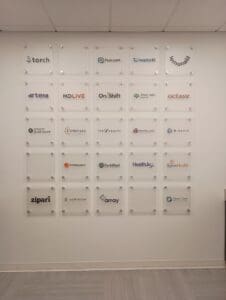 A wall featuring multiple company logos on clear plaques, displayed in a grid format inside an office.