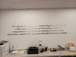 Office wall with a mission statement about generating financial returns and innovative healthcare, accompanied by a printer and a scanner on a table.