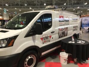 White van with ace handyman services branding parked at an indoor expo, accompanied by a promotional table with brochures and cups.