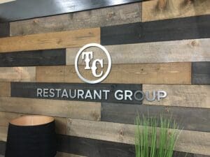 A logo of "tc restaurant group" mounted on a rustic wood-paneled wall, with varied shades of brown and black wood strips, accompanied by greenery in a pot.