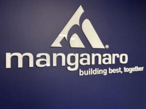 White 3d text saying "manganaro building best together" with a logo, mounted on a dark blue wall.