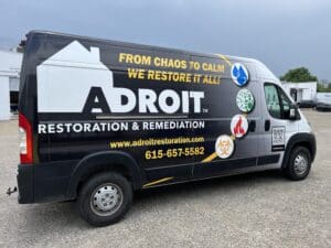 Black commercial van with "adroit restoration & remediation" company advertising, including contact details and services, parked in a lot.