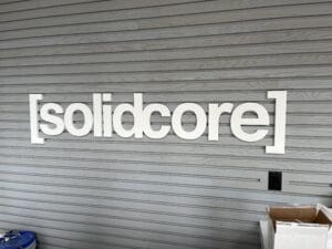 Sign reading "[solidcore]" in bold white letters mounted on a gray slatted wall, with a cardboard box and blue bin visible at the bottom.