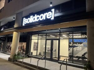 Exterior view of a [solidcore] fitness studio at night with illuminated signage, visible workout equipment inside, and promotional text on the window.