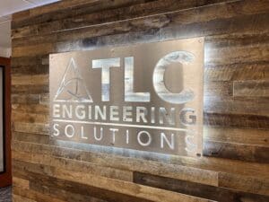 A metal sign reading "tlc engineering solutions" mounted on a wooden wall.