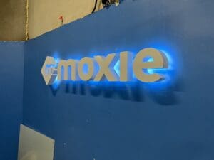 Illuminated "moxie" sign on a blue wall with sleek, modern styling and backlighting that casts a subtle glow.