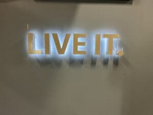 Backlit wall-mounted letters spelling "live it" casting sharp shadows on a gray wall.