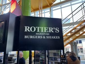 Sign for "rotier's signature burgers & shakes" hanging in a bright, modern indoor space with people and other shops visible in the background.