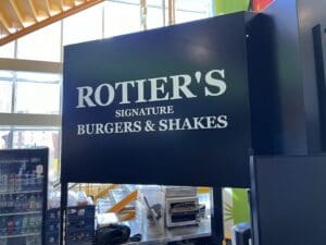 Sign overhead reading "rottier's signature burgers & shakes" in white text on a black background, visible in a brightly lit fast food setting.