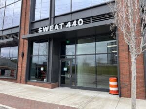 Exterior view of sweat 440 gym with black signage on a brick building, featuring glass doors and bare trees nearby.