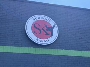 A round sign for scruggs karate on a grey wall, featuring the letter "s" with a silhouette of a karate practitioner inside the letter.