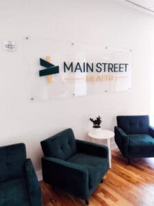 A neatly arranged waiting area with green armchairs, white side table, and a large sign reading "main street health" on a white wall.