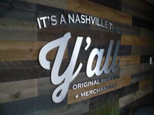 Wooden wall with the text "it's a nashville thing y'all" and "original shirt co. merchandise" in 3d silver lettering.