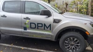 Silver pickup truck with "dpm surface care" branding on the side, parked beside a leafy curb on a wet day.