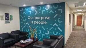 Office lobby with teal wall featuring the phrase "our purpose is people" in white text, accompanied by abstract symbols. black sofas and a coffee table with a plant and magazines are in the foreground.