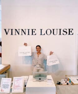 A cheerful woman standing behind a counter in a boutique, holding two shopping bags labeled "vinnie louise," with the store's name on the wall behind her.