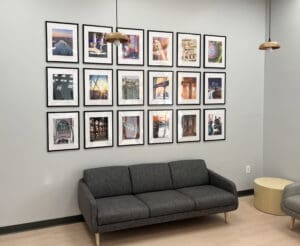 A modern waiting room with a gray sofa, a round side table, two hanging lamps, and a large gallery wall of framed travel-themed posters.