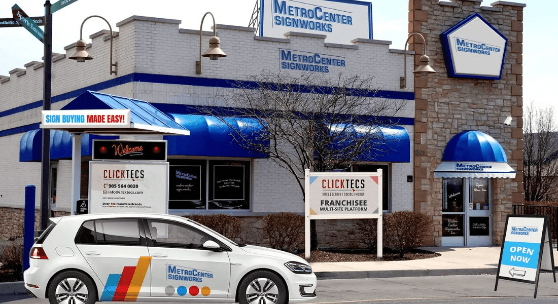 A MetroCenter Signworks shop with blue accents and a branded white car parked in front. Several signage boards are displayed around the store's exterior. The store has an "Open Now" sign.
