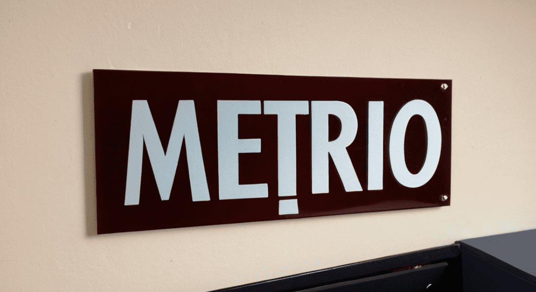 A rectangular sign with a dark red background displays the word "METRIO" in large, bold white letters. The sign is mounted on a light-colored wall. Lobby Signs: Design Tips, Types, and Benefits