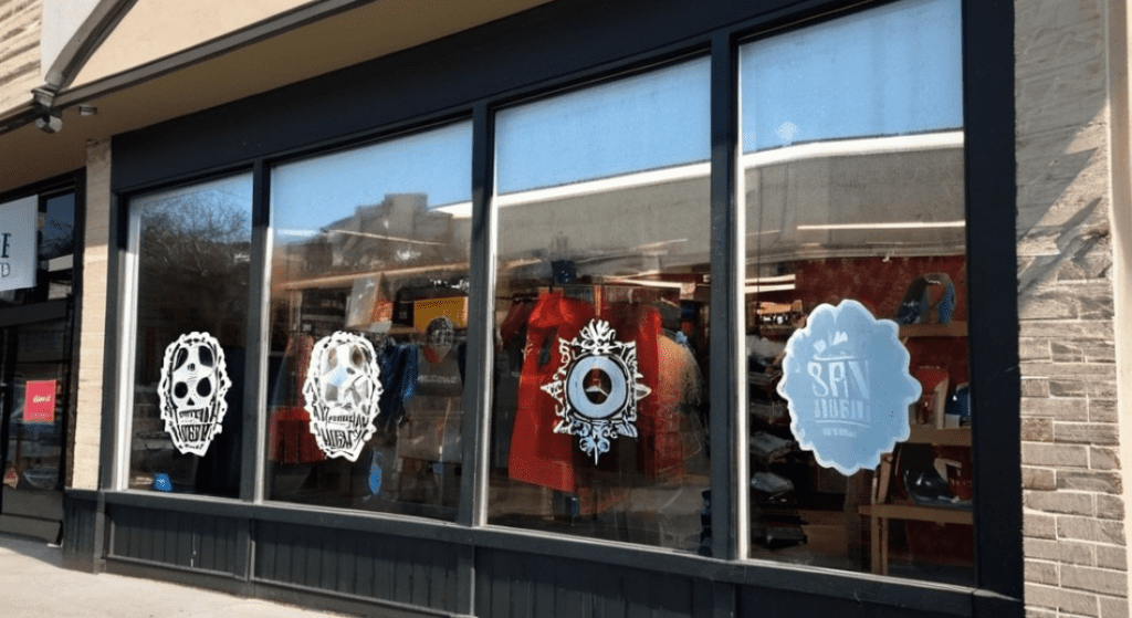 Storefront with four large windows displaying white decals and clothing items inside. Plastic snowflakes, skulls, and a circular design are visible among the decals.