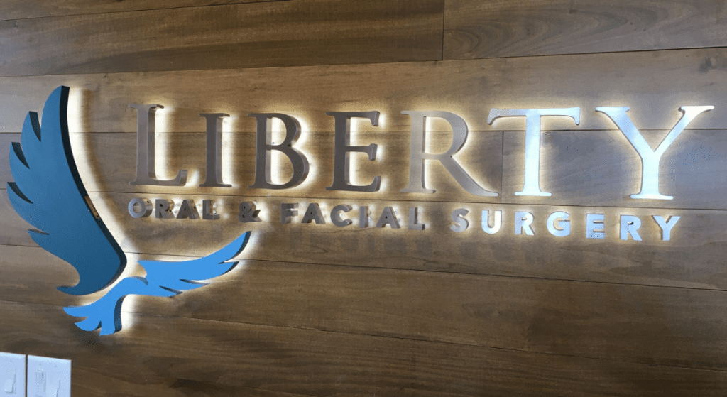 Signage for Liberty Oral & Facial Surgery on a wooden wall, featuring illuminated lettering and a blue and white abstract bird logo. Refresh Your Business Indoor Signs