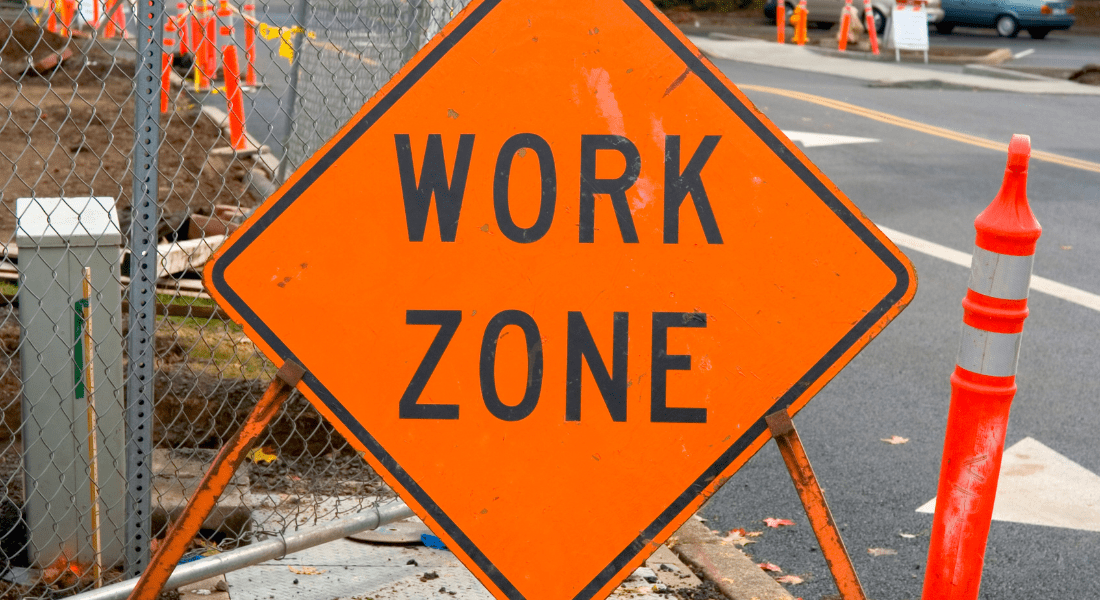 An orange "Work Zone" sign positioned at a road construction site, with visible fencing and traffic cones nearby.