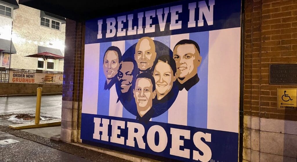 Mural on a brick wall featuring diverse group of people with text "I Believe in Heroes." Wet sidewalk in front and a Grubhub sign on the left.